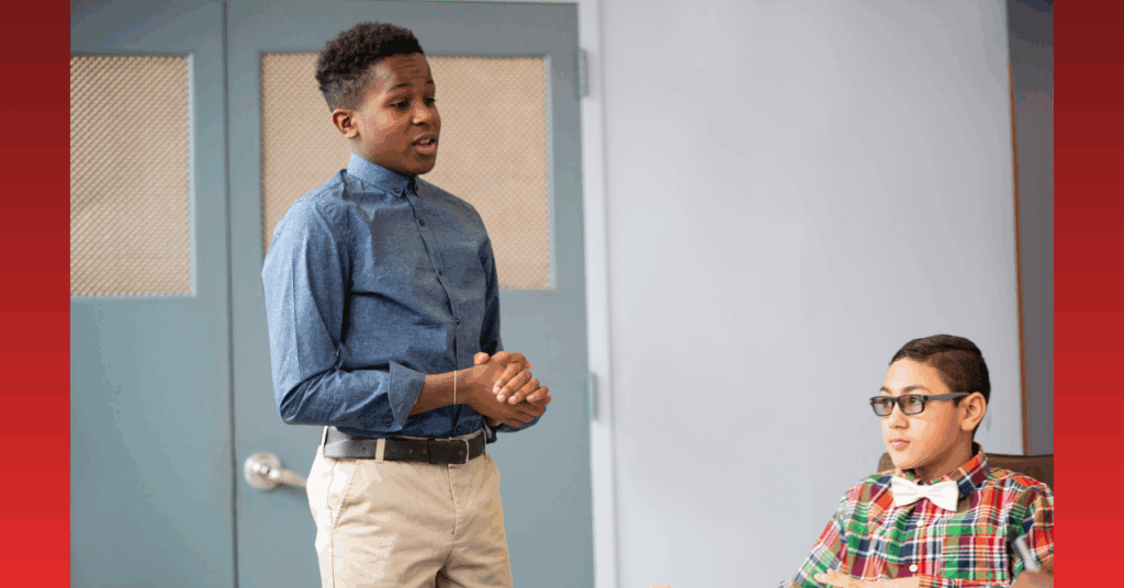 High school student giving a classroom presentation while peer listens attentively.